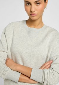 Sudadera gris claro con cuello redondo y mangas largas. La tela presenta una textura sutil y puños acanalados. El modelo tiene los brazos cruzados.