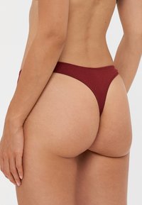 Chiloți thong burgundy cu textură nervurată. Design de tip low-rise cu bretele subțiri, care evidențiază o siluetă cu acoperire minimă la spate.