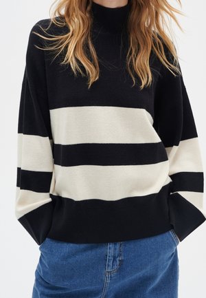 Osoba ubrana w czarno-biały, szeroko prążkowany sweter z długimi rękawami oraz niebieskie dżinsy typu denim, z rękami w kieszeniach.