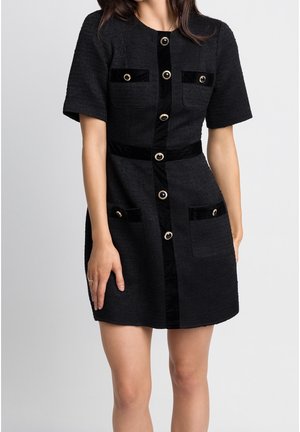 Vrouw die een zwart jurkje met korte mouwen draagt, met een voorzijde voorzien van gouden knopen en zakdetails, staand tegen een effen lichte achtergrond.