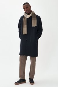 Homme portant un manteau bleu marine foncé, une écharpe rayée beige et grise, un pantalon marron clair et des baskets noires avec des semelles beige, se tenant devant un fond blanc.