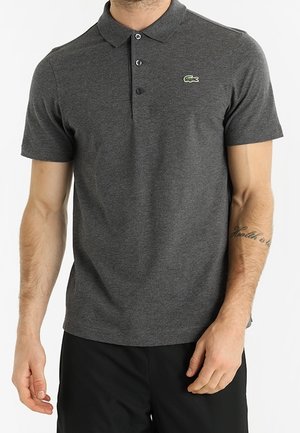 Mann trägt ein dunkles graues Polohemd mit kurzen Ärmeln, einem Zwei-Knopf-Verschluss und einem kleinen grünen Krokodil-Logo auf der linken Brust.