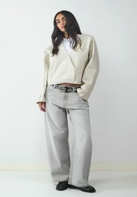 Beżowa krótka kurtka w stylu wiązanym, biała koszulka, luźne szare dżinsy, czarny pasek w kropki oraz czarne baleriny. Gładka faktura, minimalistyczny design, swobodny krój.