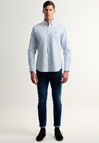Camisa de botón a rayas azul claro hecha de algodón, combinada con jeans oscuros. Cuenta con un collar y un logo bordado en el pecho.