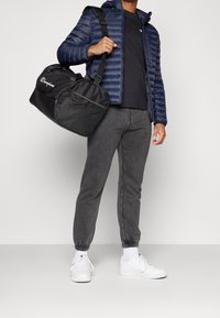 Black duffel bag with mesh accents, carried by a person in a navy puffer jacket and gray joggers, standing on a light background.