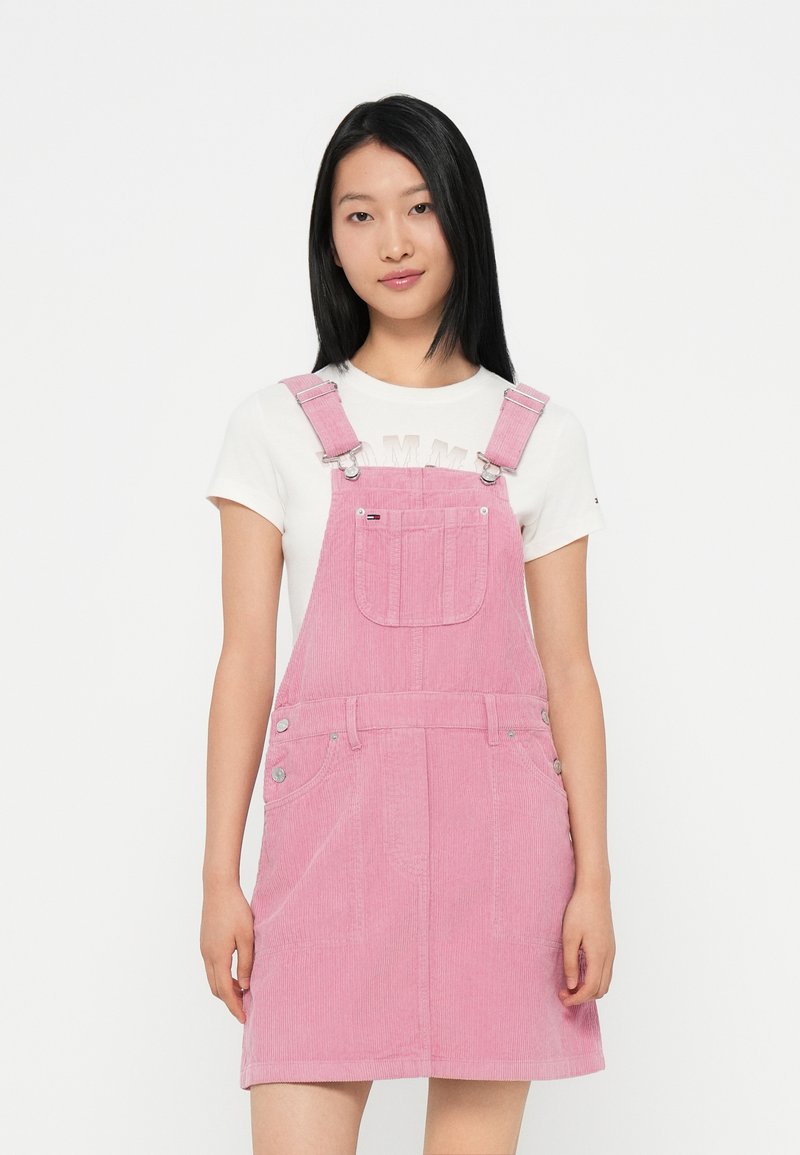 Rosa Corduroy-Latzkleid mit verstellbaren Trägern, Fronttasche und silbernen Hardware-Details. Getragen über einem weißen T-Shirt mit Logo-Druck.