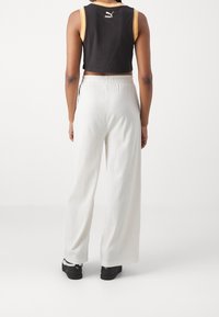 Osoba v černém crop topu Puma, bílých širokých ribbed kalhotách a černých teniskách Puma, stojí čelem k prostému bílému pozadí.