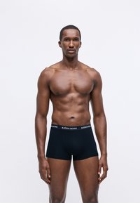 Vīrietis ar muskuļu ķermeni stāv, ģērbies melnās Björn Borg bokseršortos, ar neitrālu izteiksmi uz vienkārša balta fona.