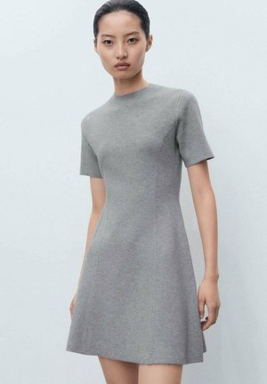 Femme debout portant une robe ajustée gris clair à manches courtes avec un col haut contre un fond clair.