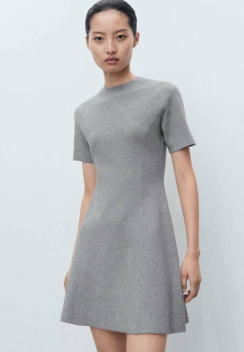 Femme debout portant une robe ajustée gris clair à manches courtes avec un col haut contre un fond clair.