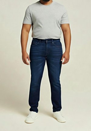 Mann trägt ein hellgraues kurzärmeliges T-Shirt, dunkelblaue enganliegende Jeans und weiße Sneaker und steht vor einem schlichten Hintergrund.