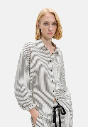 Młoda kobieta z krótkimi blond włosami, ubrana w luźną białą koszulę na guziki oraz pasujące do niej spodnie w paski z czarnym sznurkiem.