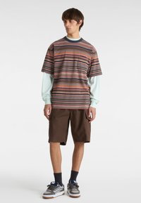 Camiseta de rayas con mangas cortas, que presenta rayas verticales multicolores. Llevada sobre una camisa de manga larga azul clara y combinada con shorts marrones.