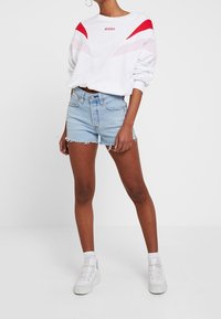 Vit sweatshirt med röda och rosa geometriska detaljer och en logotyp. Ett par ljusblå denimshorts med fransad nederkant. Vita sneakers.