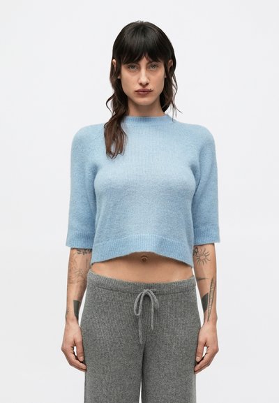Pull court bleu clair à manches courtes, confectionné dans un tissu doux. Porté avec un pantalon gris à cordon de serrage, présentant une texture côtelée.