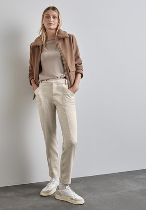 Brun zip-up jacka med plyschkrage, beige topp, ljusa strukturerade byxor och vita sneakers med grå detaljer, mot en neutral bakgrund.