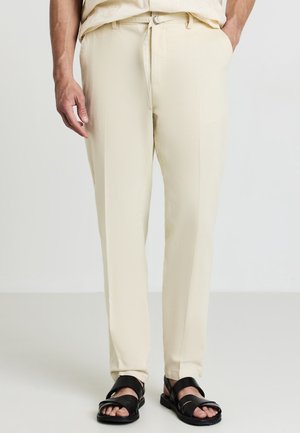 Pantaloni dritti color beige chiaro con una finitura testurizzata, vita con lacci e tasche laterali, abbinati a sandali neri con cinturini.