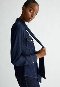 Giacca di jeans blu scuro con due tasche sul petto, bottoni argentati e cuciture a contrasto. Il tessuto ha una texture liscia e un taglio strutturato.