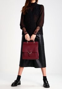 Sac à dos en cuir bordeaux avec un rabat et un détail anneau, forme rectangulaire, poche avant plate, associé à un haut en dentelle noire et une jupe en cuir.