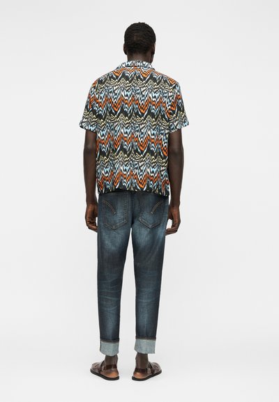 Άνδρας που φοράει πολύχρωμο πουκάμισο με μοτίβο ζιγκ-ζαγκ και κοντά μανίκια, σκούρα τζιν με σηκωμένα μπατζάκια και καφέ σανδάλια, στέκεται γυρισμένος προς τα πίσω σε λευκό φόντο.