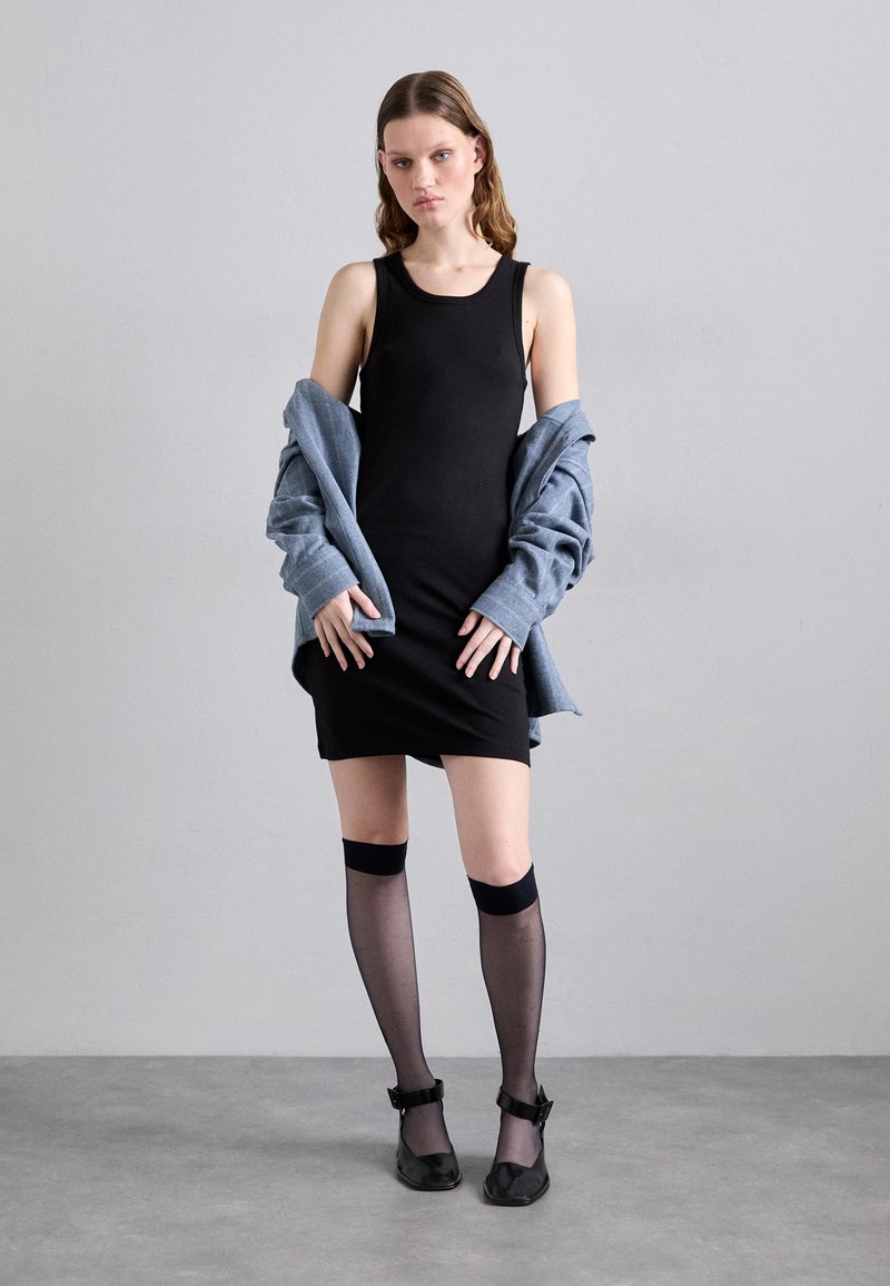 Robe noire sans manches, grosse veste bleu clair drapée sur les épaules, collants noirs transparents jusqu'aux genoux et chaussures à bride de cheville noires.