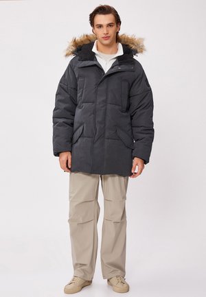 Parka noire matelassée avec une capuche doublée de fourrure, dotée de multiples poches avant et d'une fermeture éclair. Portée avec un pantalon beige et des baskets claires.