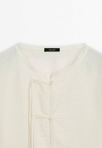 Chemise de couleur crème avec encolure ronde, deux petits boutons et fines boucles de tissu le long du centre avant.