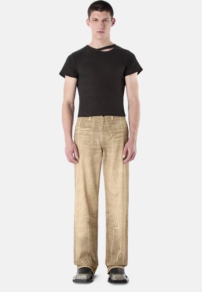 Homme debout portant un T-shirt noir ajusté avec découpe aux épaules, un pantalon large à carreaux beige, et des chaussures noires à bout carré sur fond blanc.