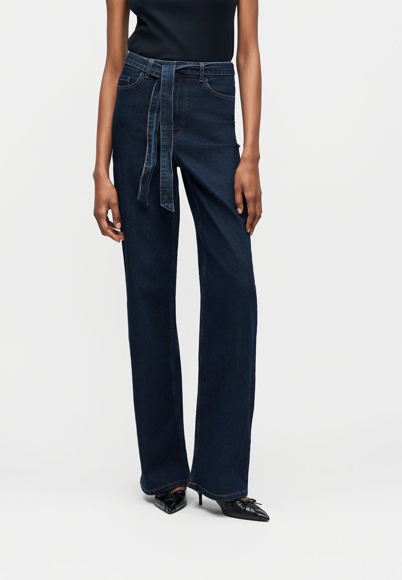 Γυναίκα που φοράει σκούρο μπλε τζιν με ψηλή μέση και άνετη γραμμή, συνοδευόμενο από ζώνη από το ίδιο ύφασμα και μαύρες μυτερές γόβες.