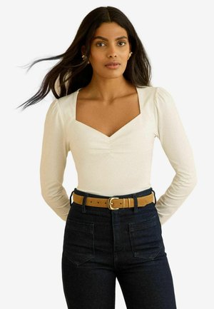 Cremefarbenes langärmeliges Bodysuit mit quadratischem Ausschnitt und gerafftem Detail, kombiniert mit hoch taillierter dunkelblauer Jeans und einem braunen Gürtel.