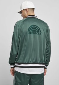 Giacca bomber verde in satin con colletto e polsini a strisce bianche e nere. Presenta un grande logo nero ad arco con il testo "SOUTHPole" sulla schiena.