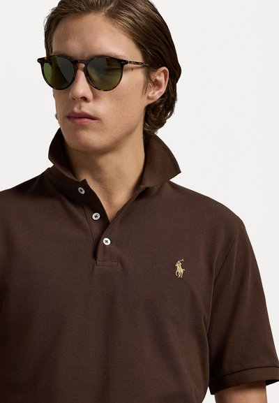 Polo marron avec un col rehaussé, une patte de boutonnage à trois boutons et un logo brodé doré. Les lunettes de soleil ont une monture en écaille de tortue et des verres verts.