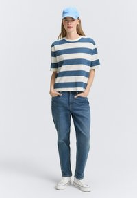 Gestreiftes T-Shirt in Creme und Marineblau, lockere Passform; hellblaues Cap; klassische blaue Jeans; weiße Sneaker mit schwarzen Akzenten.