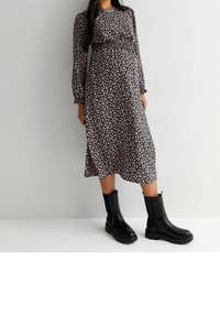 Musta kukallinen äitiysmekko, jossa on pitkät hihat ja istuva bodice. Yhdistetty polven yli ulottuviin mustiin nahkasaappaisiin, joissa on paksu pohja.
