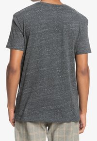 Persona con una camiseta de manga corta de color gris oscuro jaspeado y pantalones de cuadros beige, vista desde atrás contra un fondo blanco.