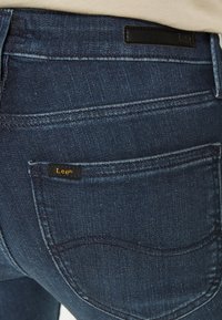 Tummansiniset farkut, joissa on musta nahkatalvi, näkyvät saumat, klassinen taskusuunnittelu ja pieni logotagi.