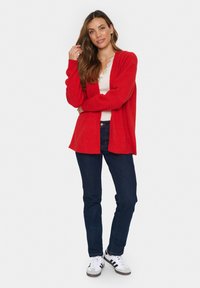 Rode gebreide cardigan met lange mouwen en een open voorkant, gecombineerd met een witte top en donkere jeans. Draag het met witte en zwarte sneakers.