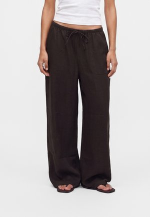 Femme portant un pantalon large noir ample avec poches avant et taille à cordon, associé à des tongs noires et un top court blanc.