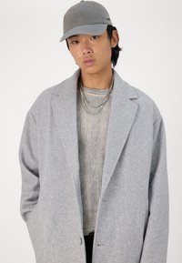 Cappotto in lana oversize grigio chiaro con ampi revers, abbinato a un maglione grigio a coste e catene d'argento sovrapposte. Il modello indossa un cappello grigio.