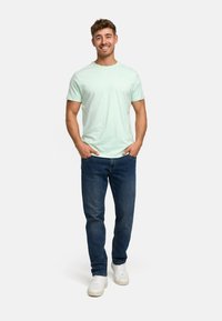 Light green cotton t-shirt with short sleeves, paired with dark blue denim jeans and white sneakers. Model poses with hands in pockets.