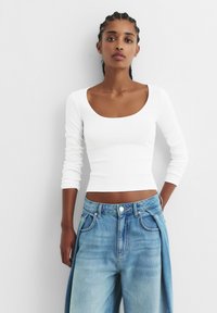 Haut blanc à manches longues côtelées avec un décolleté en scoop, associé à un jean en denim bleu clair avec une coupe ample et une texture délavée.