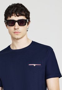 Maglietta blu navy a maniche corte con una piccolo taschino decorato da una striscia rossa e bianca, indossata con occhiali da sole neri e spessi.
