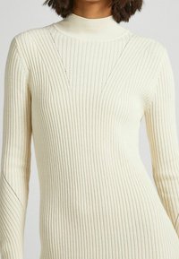 Stickad ribbad klänning i krämfärgad, med hög krage, långa ärmar och subtila triangulära utskärningar längs överkroppen.