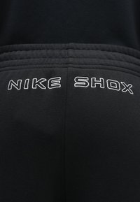 Czarne sportowe spodnie z haftowanym na biało napisem "NIKE SHOX" z tyłu, wykonane z miękkiego, teksturowanego materiału z elastycznym pasem.