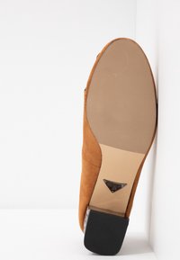 Semelle d'une mule en daim marron avec une zone de grip beige texturée et un talon bloc noir reposant contre un mur blanc.