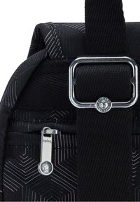 Gros plan d’un sac en tissu noir avec un curseur de fermeture éclair argenté et une boucle métallique sur une bandoulière réglable, présentant un motif géométrique et le logo Kipling.