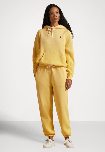 Rumena flis kapuca z žepom spredaj in belimi vrvicami, združena z ujemajočimi se ozkimi trenirkami. Model nosi bež superge.