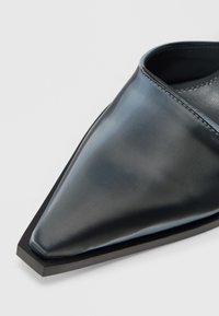 Μαύρο παπούτσι με μυτερή μύτη, κατασκευασμένο από λείο, γυαλιστερό υλικό, με αιχμηρό, γωνιώδες σχήμα και αντίθετη ματ μαύρη σόλα.