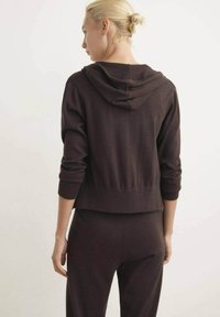 Donna bionda con i capelli legati, indossa un hoodie marrone scuro e dei pantaloni coordinati, in piedi di spalle su uno sfondo neutro.