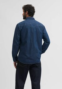Tumšas denim krekls ar garām piedurknēm, pogu aprocēm un aizmugures yoke detaļu. Iezīmēts ar norādītu apkakli un taisnu apakšmalu.